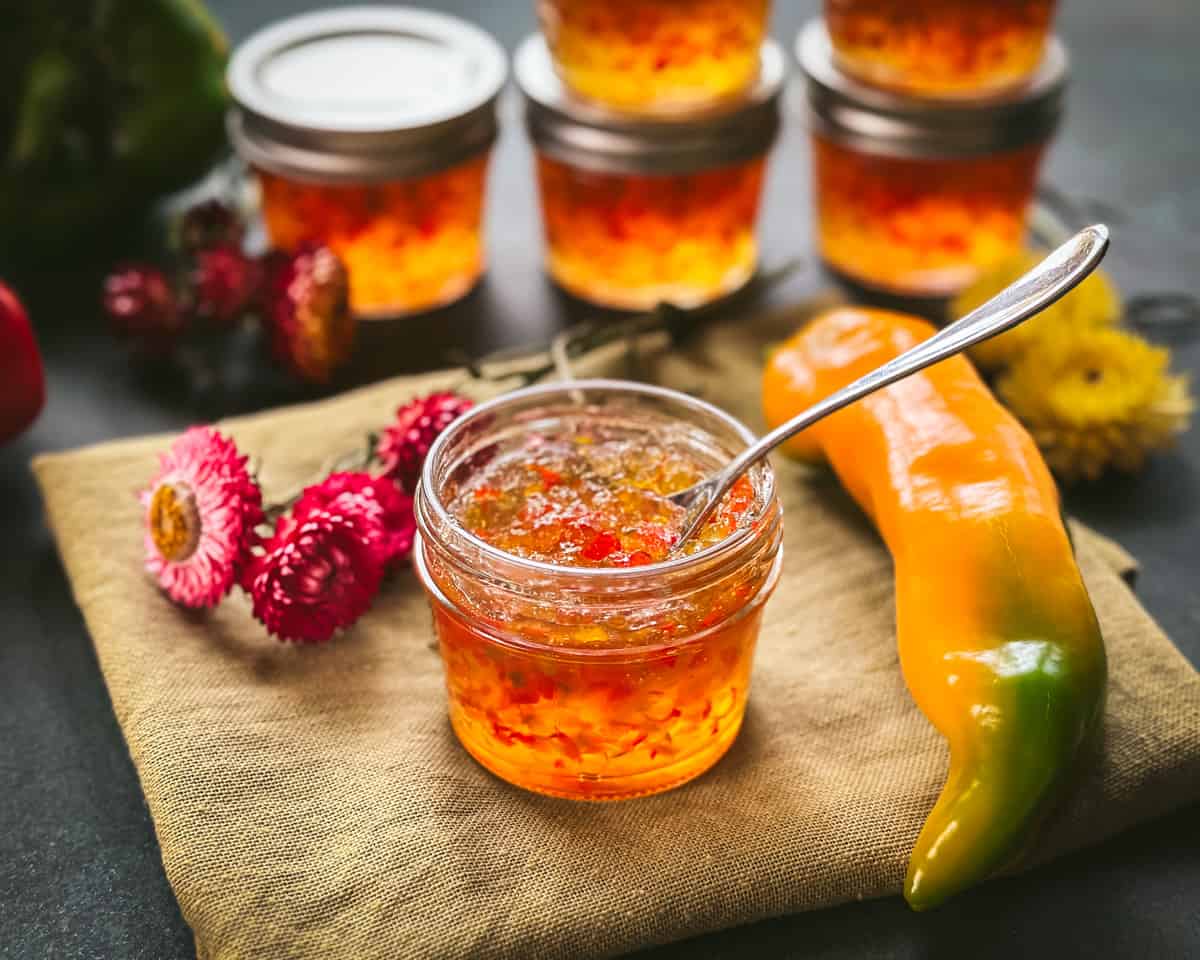 A jar of pepper jelly opened with a spoon resting in it, on a wood cutting board surrounded by flowers and a long homegrown orange bell pepper. 