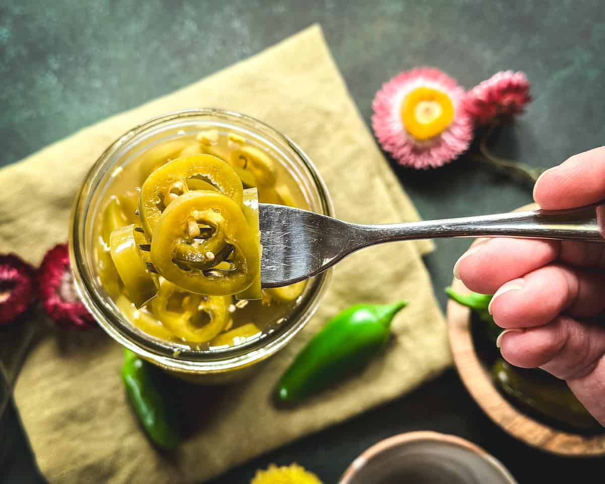 A jar of pickled jalapeños with a fork holding one up, surrounded by fresh peppers and flowers, top view.