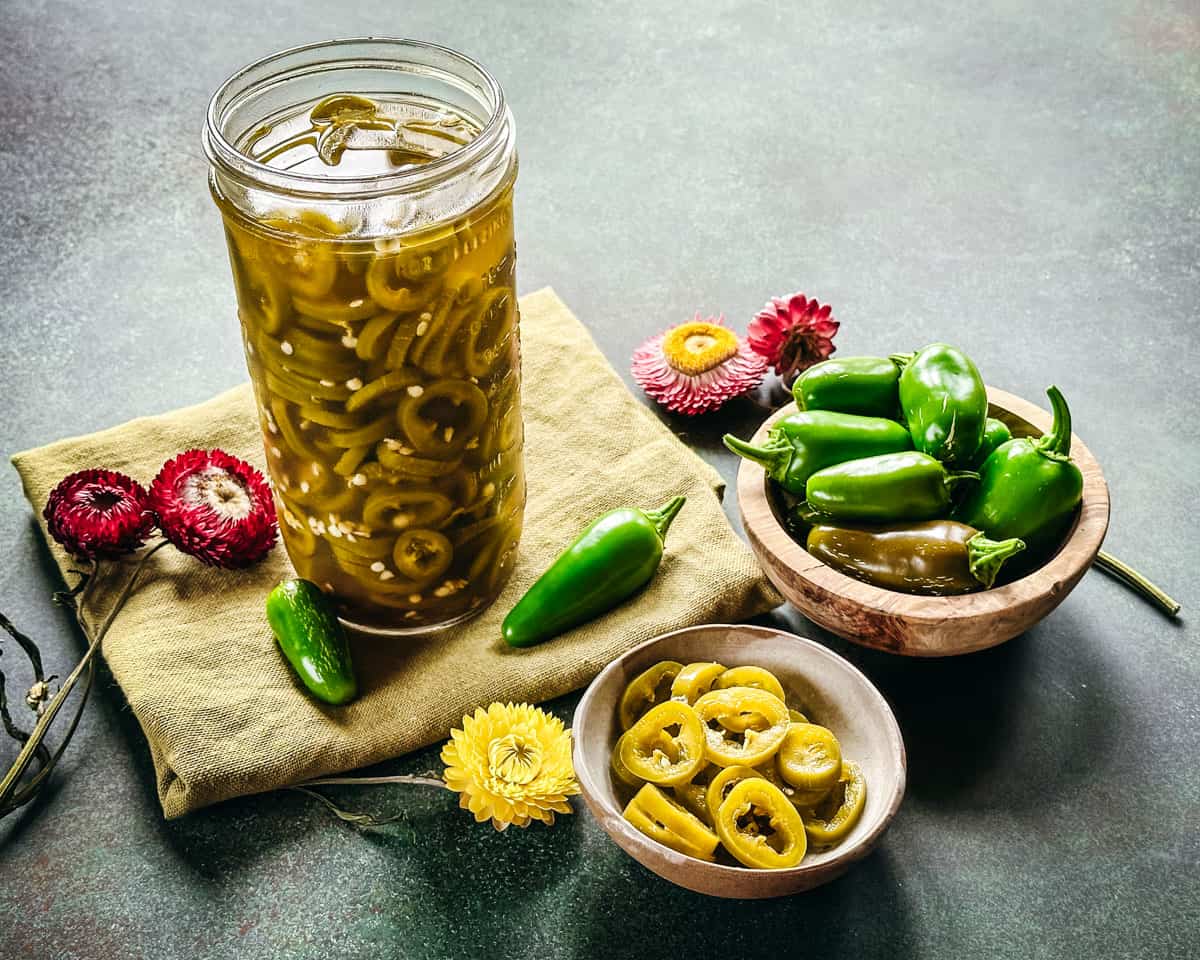 Pickled jalapeños in an opened jar on a cloth napkin, surrounded by bowls of pickled jalapeños, fresh jalapeños, and flowers. 