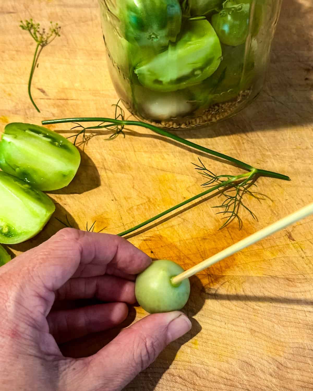 A green cherry tomato being poked with a bamboo skewer on a wood cutting board surrounded by wedged green tomatoes. 