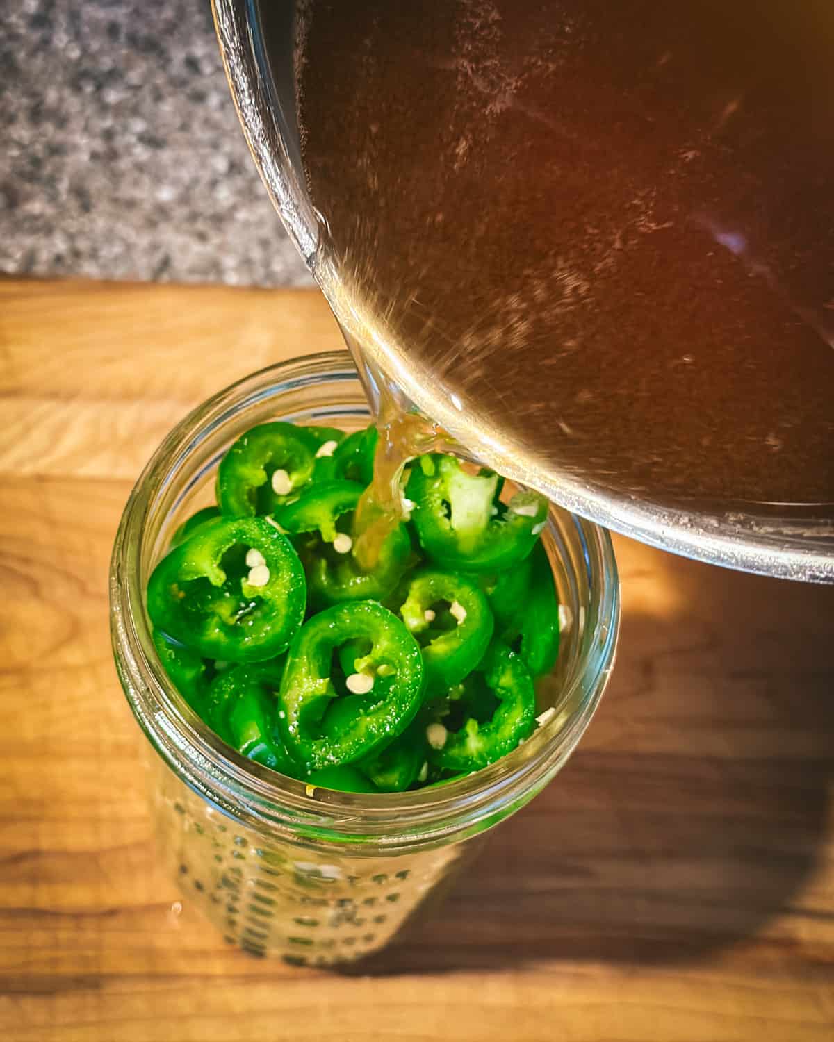 A pot of brine pouring into a jar filled with sliced jalapeños on a wood cutting board, top view. 