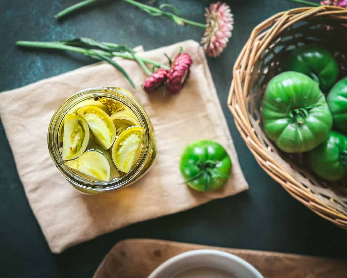 A jar of pickled green tomatoes on a wood cutting board next to a basket of green tomatoes and a burgundy flower.