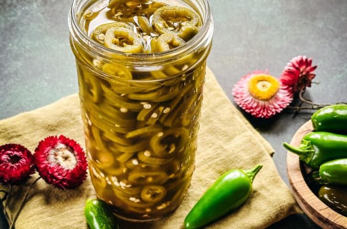 A jar of pickled jalapeño peppers on a cloth napkin, surrounded by fresh jalapeños and flowers.