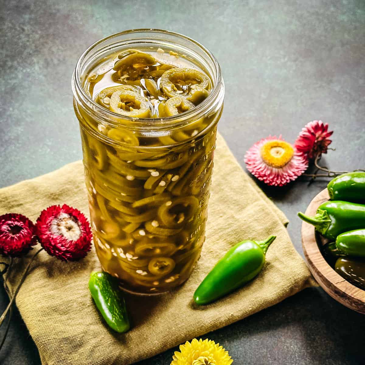 A jar of pickled jalapeño peppers on a cloth napkin, surrounded by fresh jalapeños and flowers. 