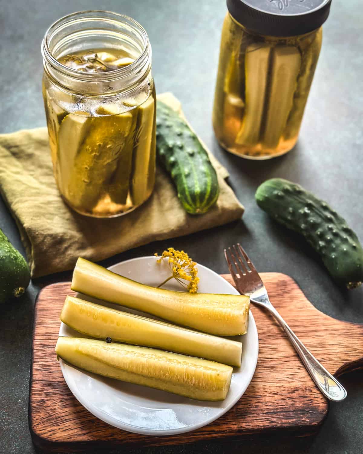 A finished jar of pickles opened with some pickle spears laying on a plate with a fork beside it on a wooden cutting board. Surrounded by a closed jar of pickles and fresh cucumbers.