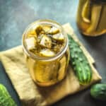 A jar of pickles on a natural cloth napkin, with fresh cucumbers surrounding.