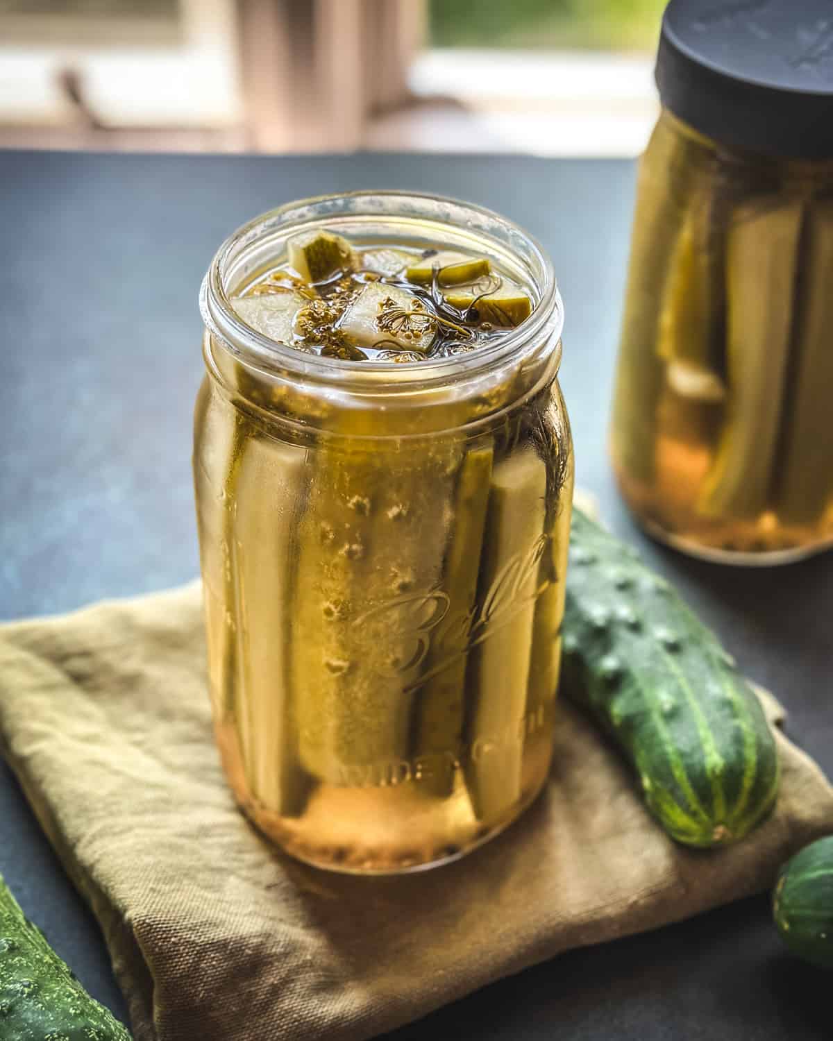 A jar of pickles on a natural cloth napkin, with fresh cucumbers surrounding.
