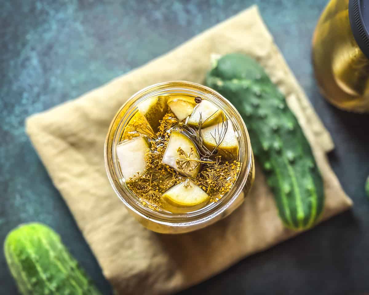 A jar of pickles on a natural cloth napkin, with fresh cucumbers surrounding, top view.