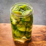A jar of pickled green tomatoes with lid off, on a wood cutting board.