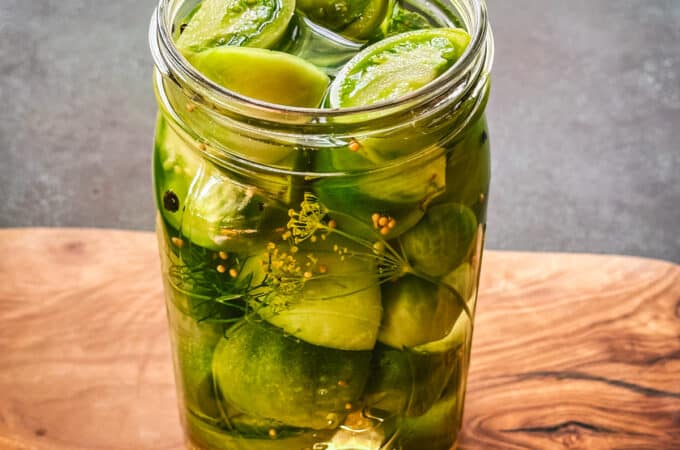 A jar of pickled green tomatoes with lid off, on a wood cutting board.