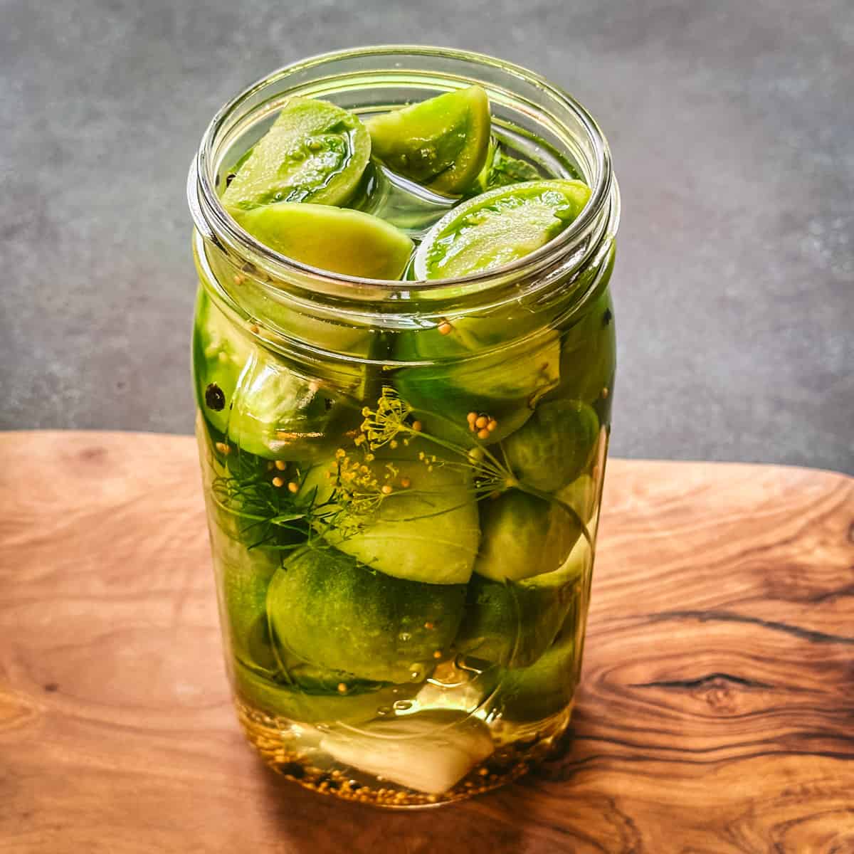 A jar of pickled green tomatoes with lid off, on a wood cutting board. 