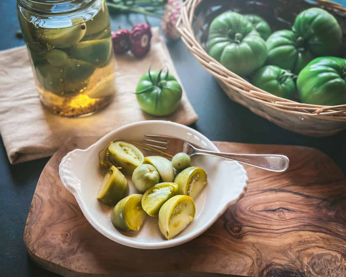 Pickled green tomatoes in a white dish with a fork, on a wood cutting board, surrounded by a jar of pickled tomatoes and a basked of green tomatoes. 