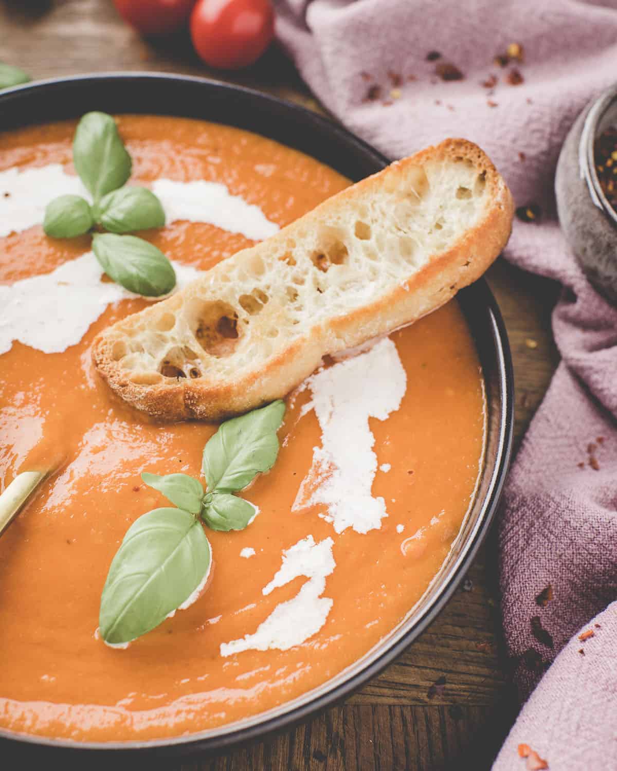 Close up of a bowl of roasted tomato soup with a swirl of cream and topped with fresh basil leaves and a slice of fresh bread. 