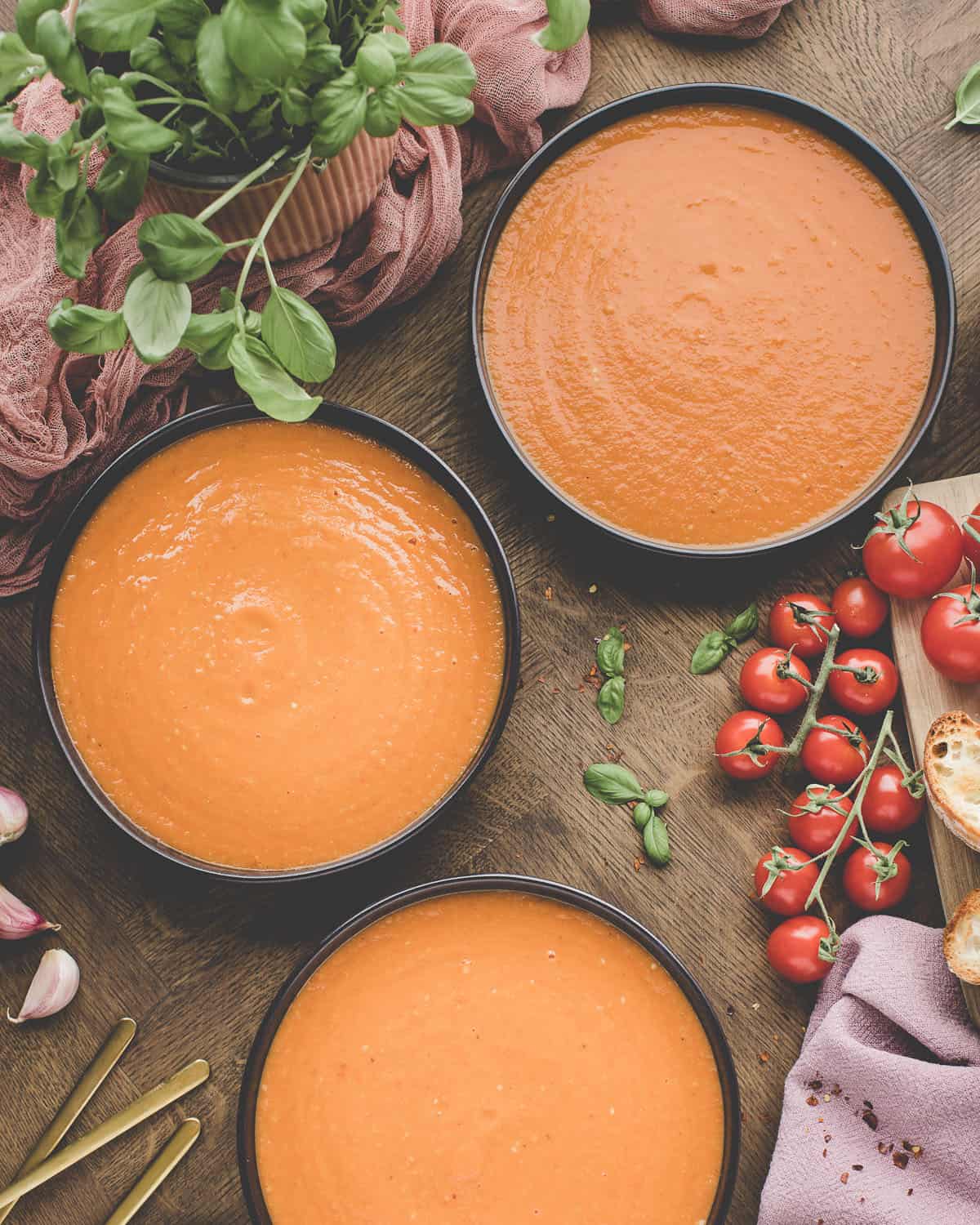 3 black bowls full with roasted tomato soup, on a wood surface surrounded by fresh tomatoes and basil. 