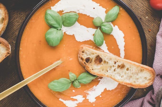 A black bowl of tomato soup with a swirl of cream, fresh basil leaves on top, and a slice of fresh bread on the edge, top view.