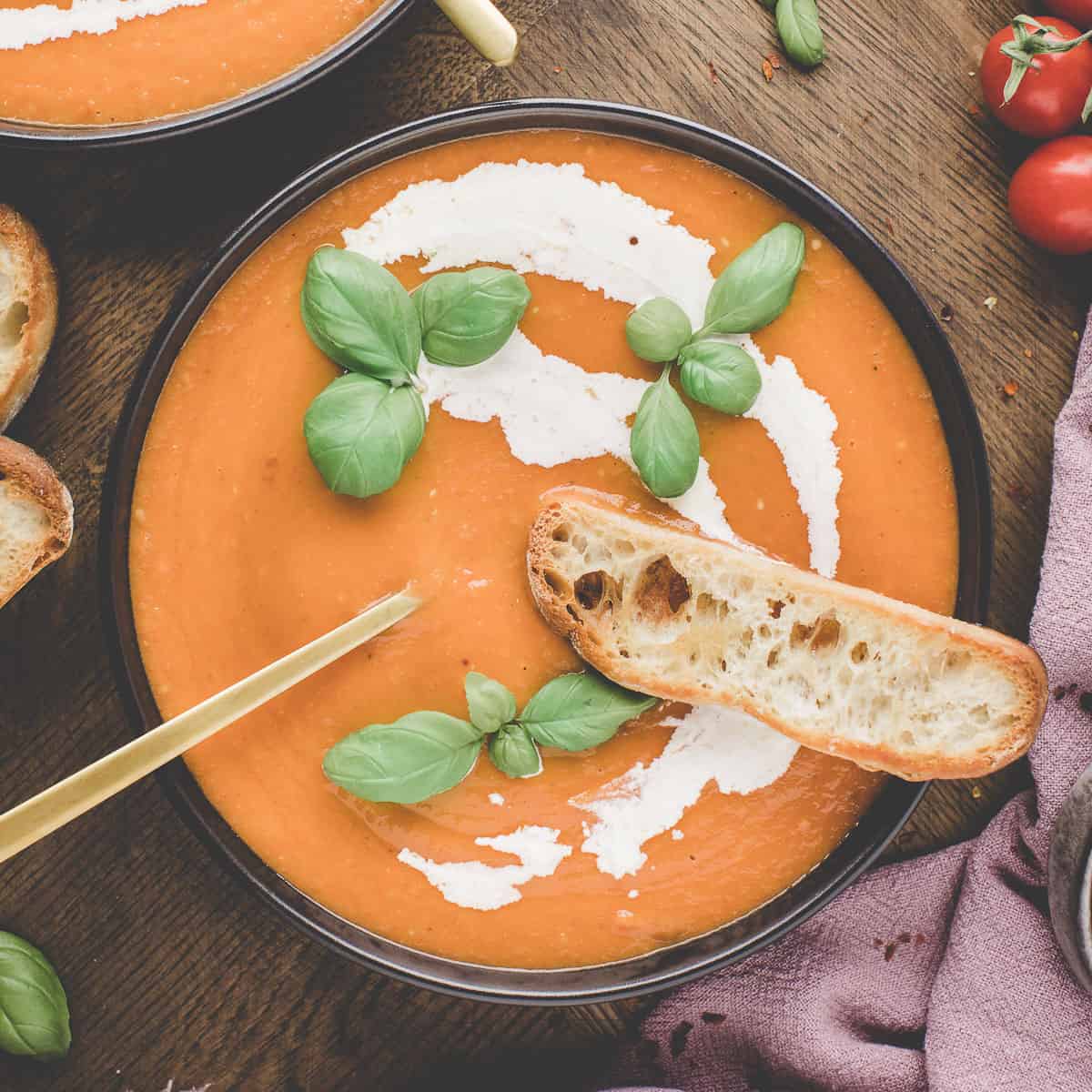 A black bowl of tomato soup with a swirl of cream, fresh basil leaves on top, and a slice of fresh bread on the edge, top view. 