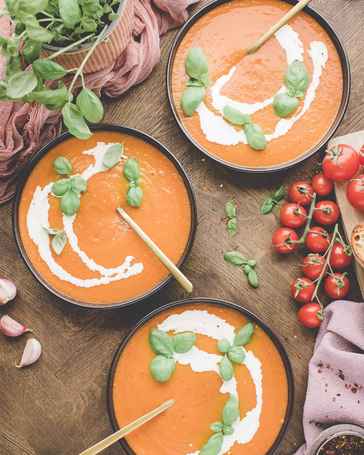 3 bowls of tomato soup with swirls of heavy cream topped with fresh basil leaves, surrounded by fresh tomatoes and basil, on a wood surface. Top view.