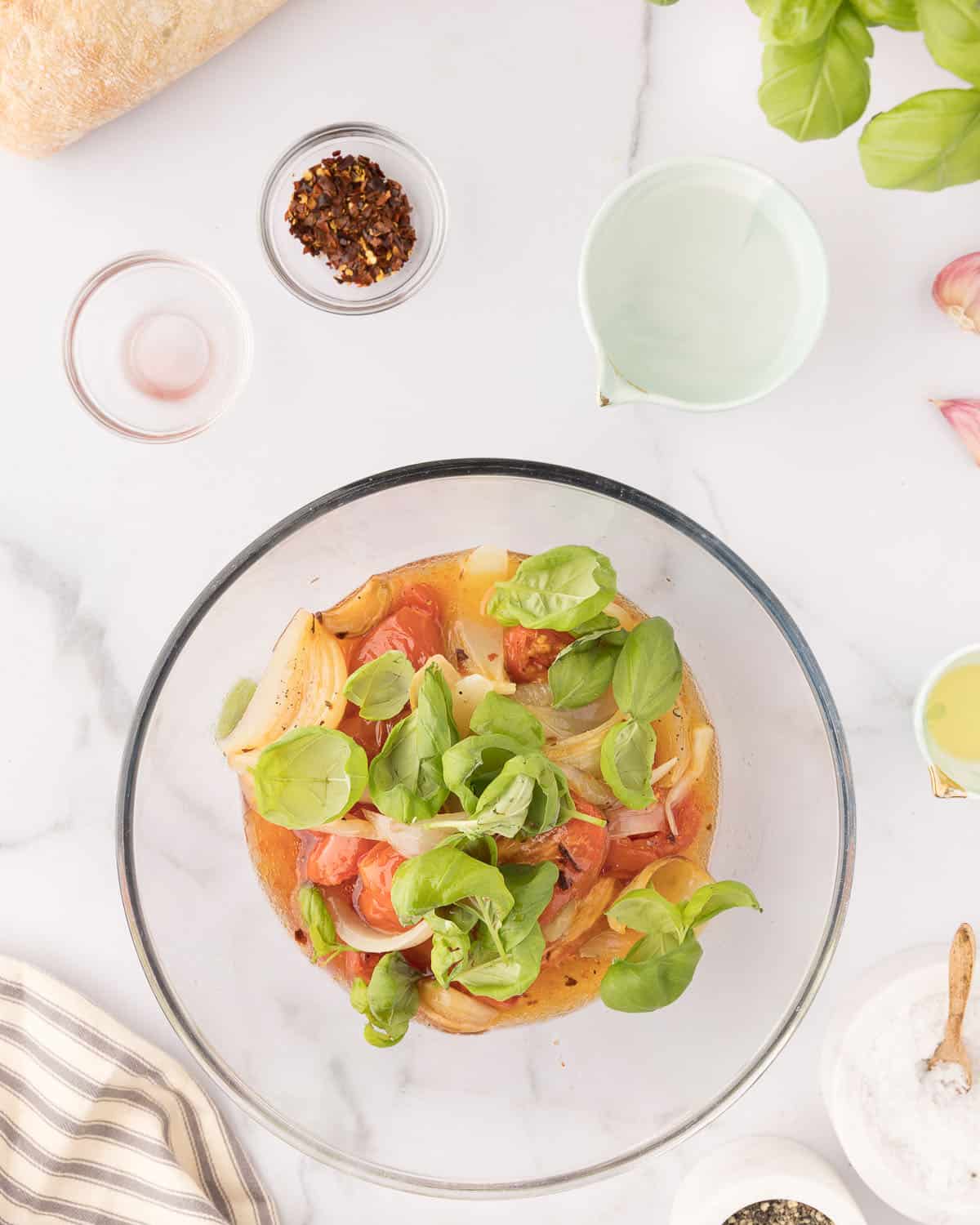 A clear bowl with the roasted vegetables in it, along with fresh basil and surrounded by small bowls of other ingredients. 