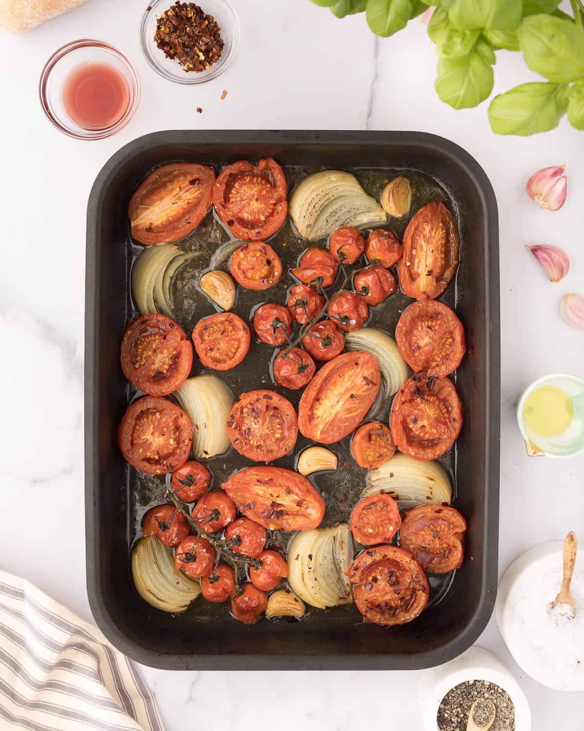 Roasted tomatoes, onion wedges, and garlic cloves in a pan, with other ingredients surrounding in small bowls, top view.