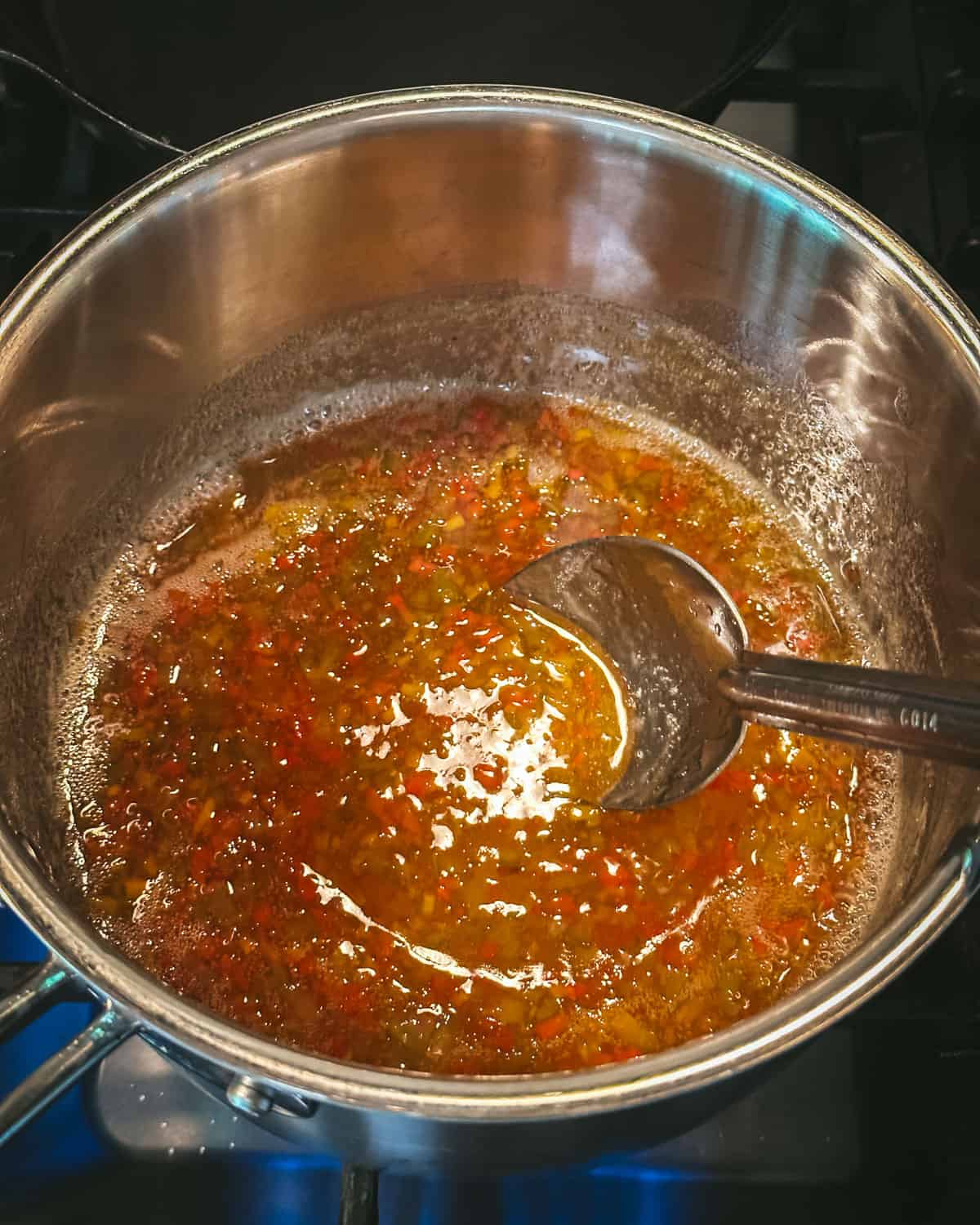 Pepper jelly cooking in a pot and being stirred with a long handled spoon. 
