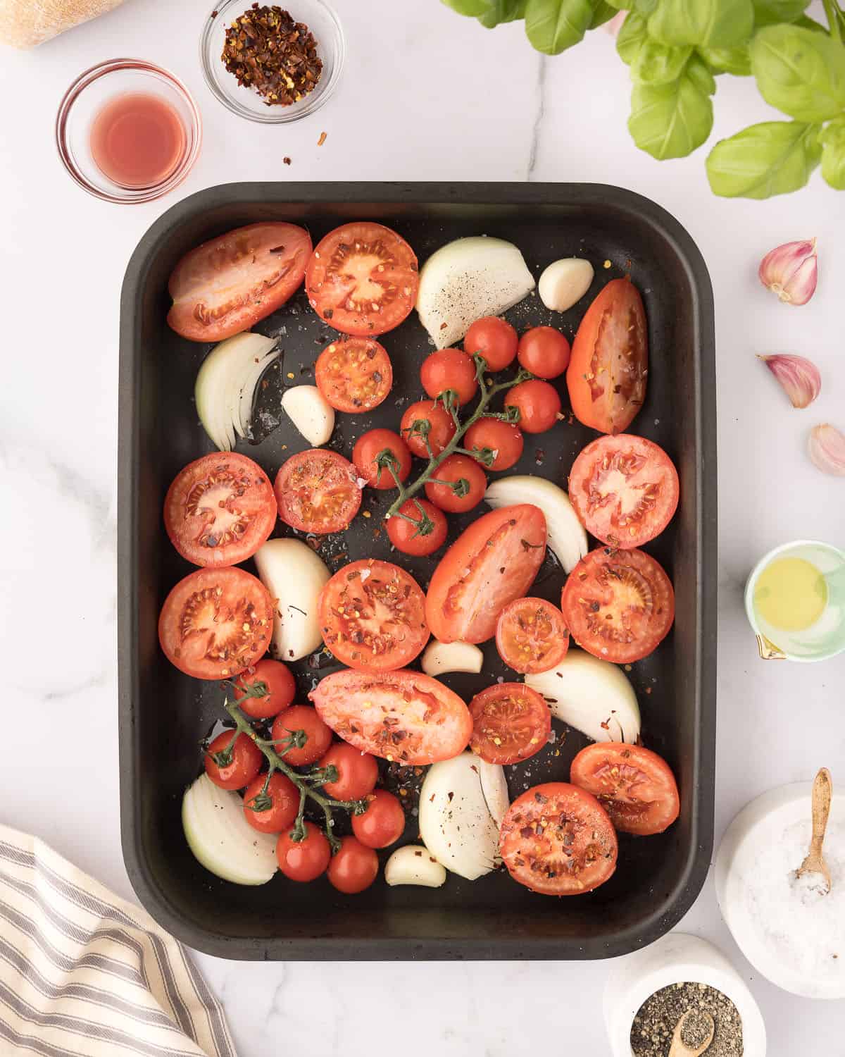 Tomatoes, onion wedges, and whole garlic cloves arranged in a baking an, surrounded by small bowls of olive oil, vinegar, and whole garlic cloves. Top view.