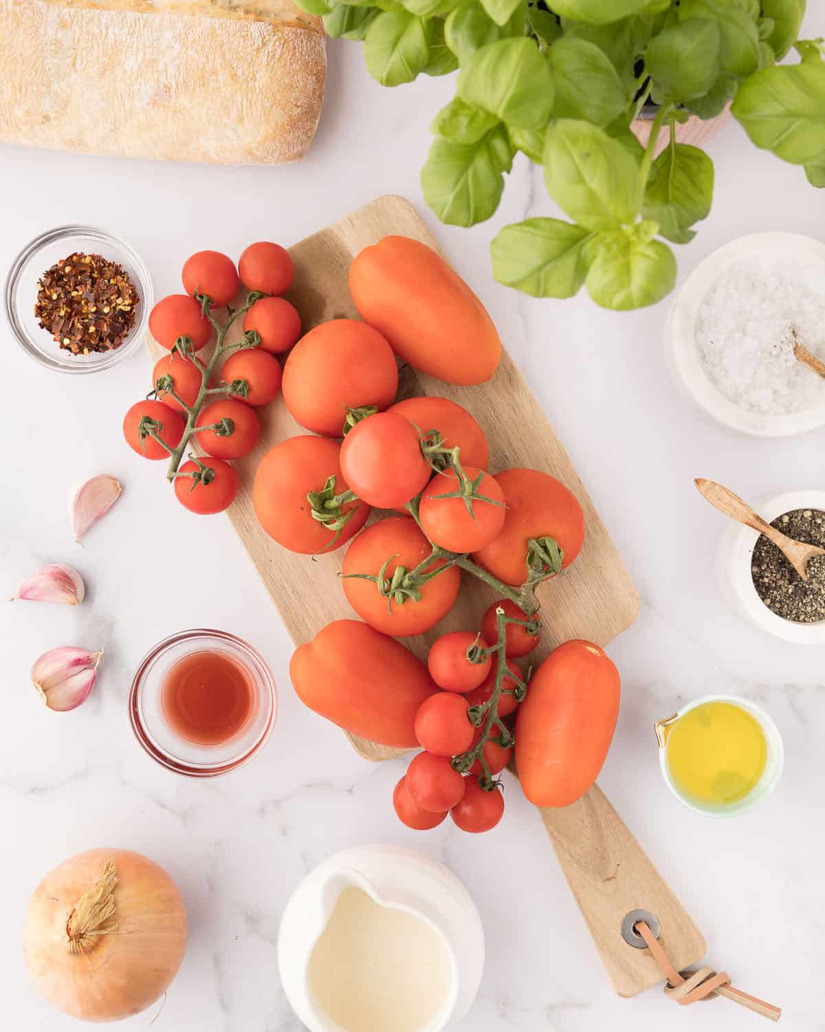 Fresh tomatoes of different sizes on a wood cutting board set on a white countertop, with other ingredients surrounding in small bowls, along with garlic cloves and fresh basil.