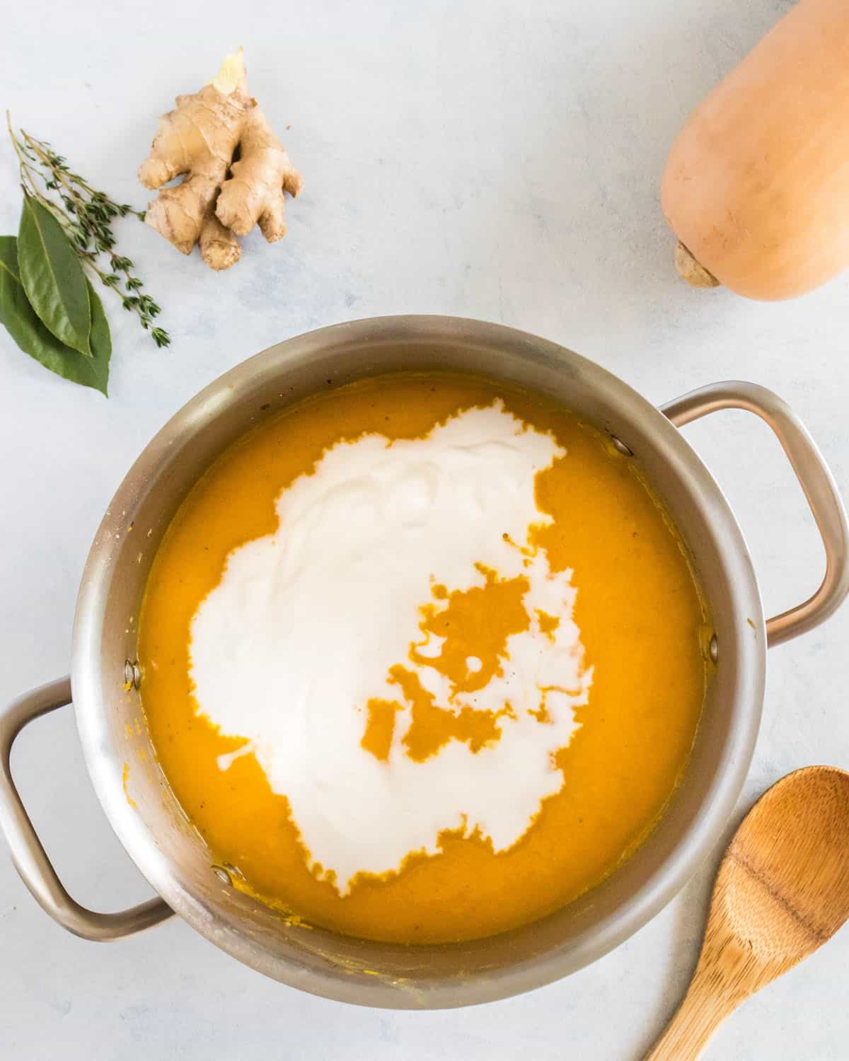 Soup pot with blended butternut squash soup drizzled with heavy cream, with a wooden spoon, a whole butternut squash, and fresh herbs surrounding. Top view. 