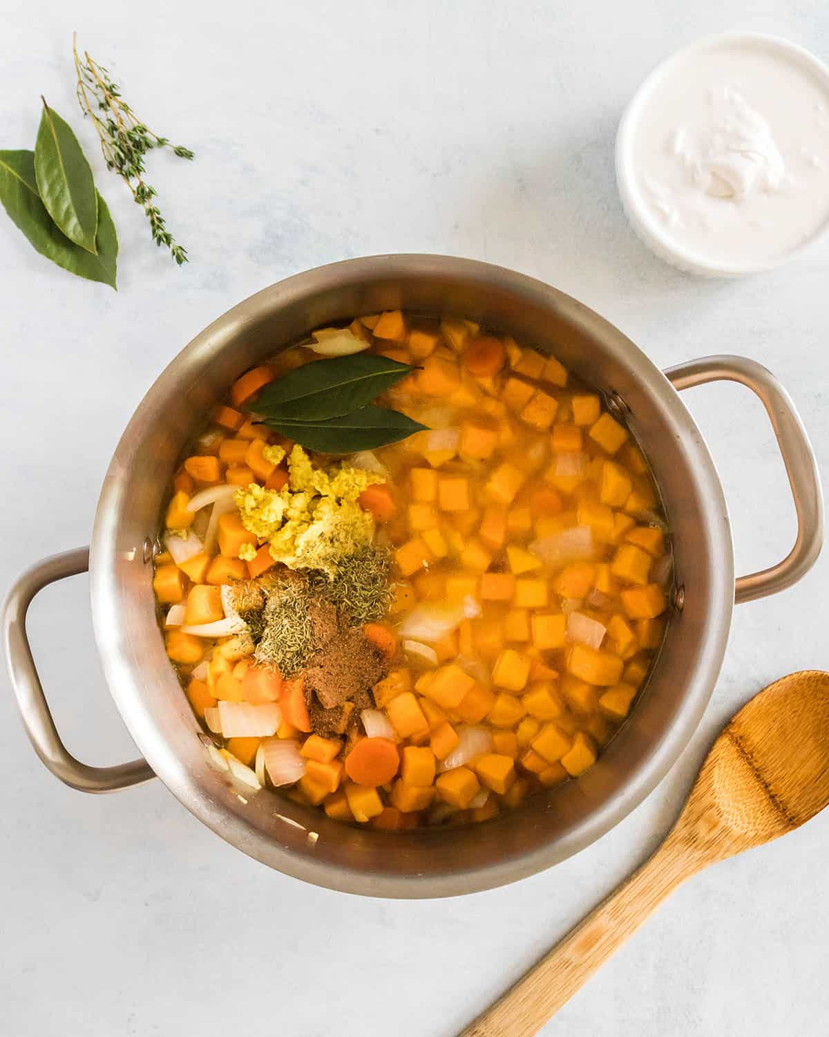 Soup pot with cooked vegetables and butternut squash cubes covered with broth, with a wooden spoon and a bowl of coconut milk to the side. 