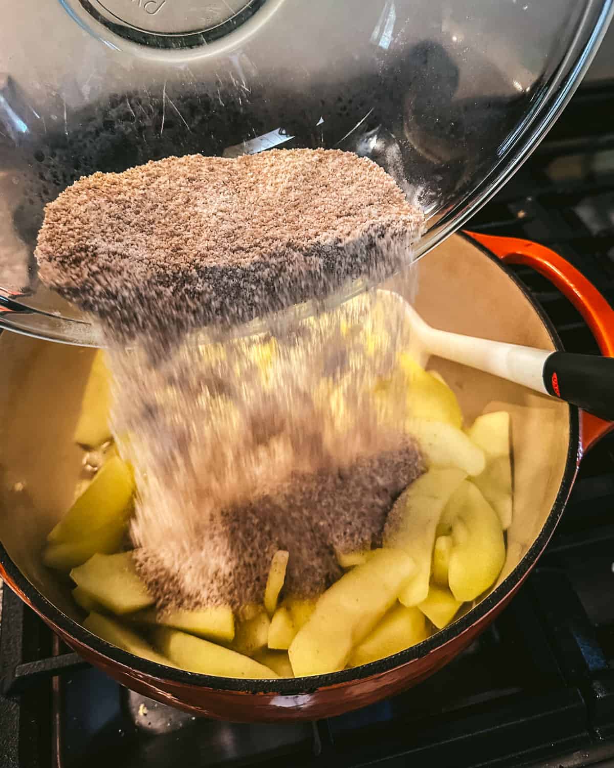 Spice mixture pouring from a clear bowl into the pot with sliced apples. 