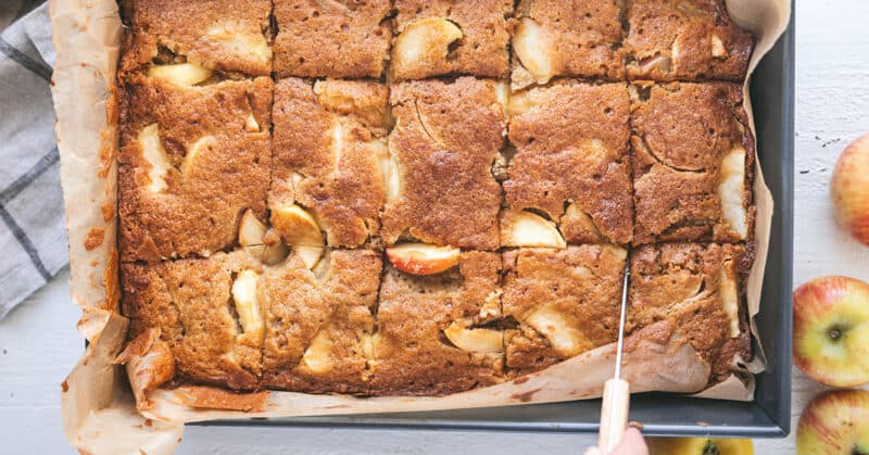 A baked apple cake in a parchment lined pan being cut into squares.