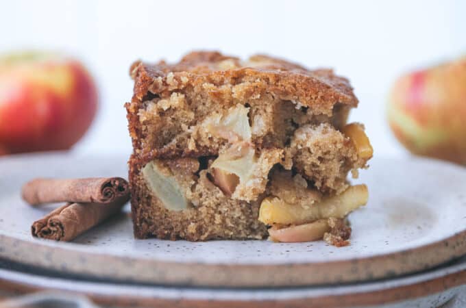 A piece of apple cinnamon cake on a white plate with whole cinnamon sticks on the plate too. Side view, with whole apples in the background.