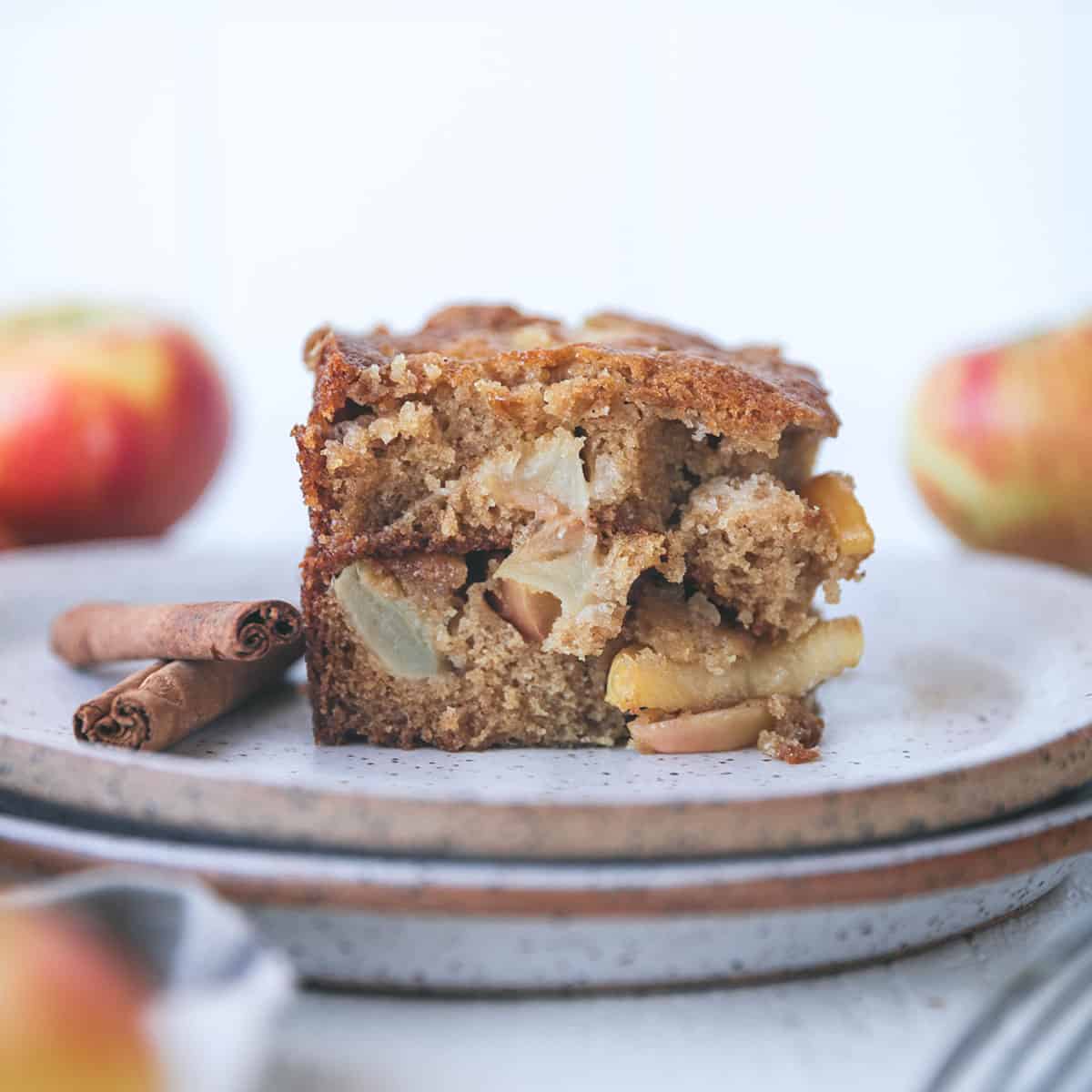 A piece of apple cinnamon cake on a white plate with whole cinnamon sticks on the plate too. Side view, with whole apples in the background. 