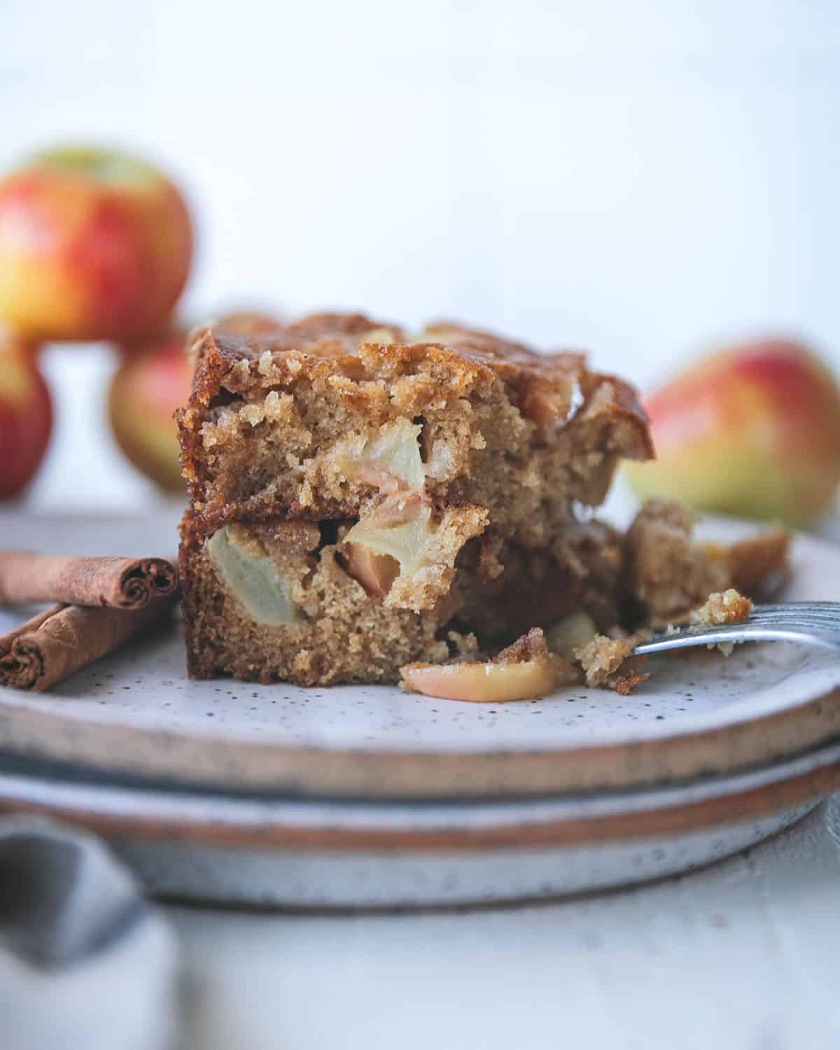 A square of apple cake on a plate, showing the side view with apple slices showing in the cake, cut into with a fork. Whole apples in the background. 