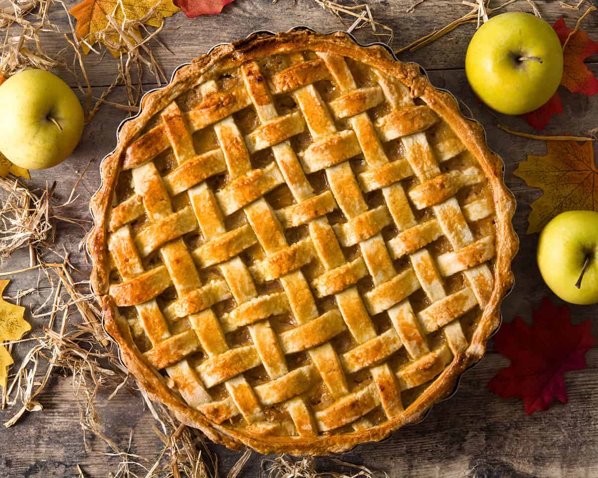 apple pie with a lattice top on a wooden table surrounded by apples and fall leaves