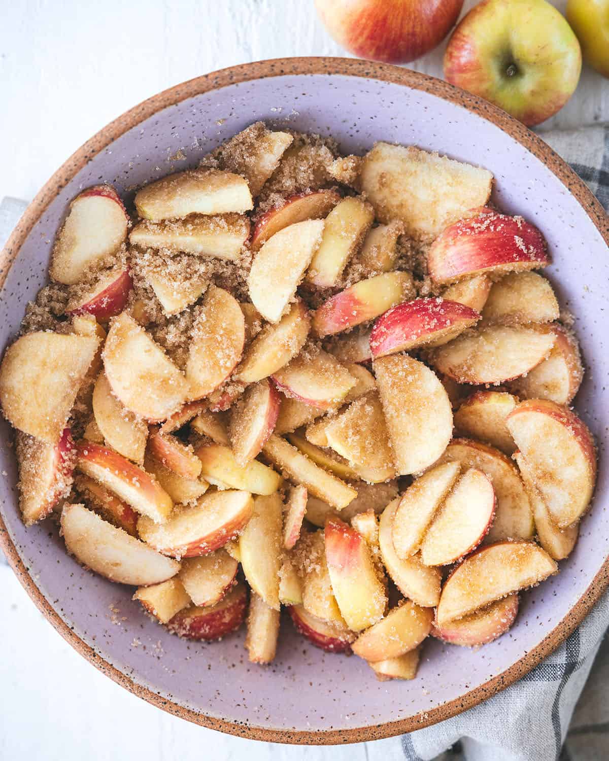 Apple slices with sugar and brown sugar mixed in a large white bowl, top view. 