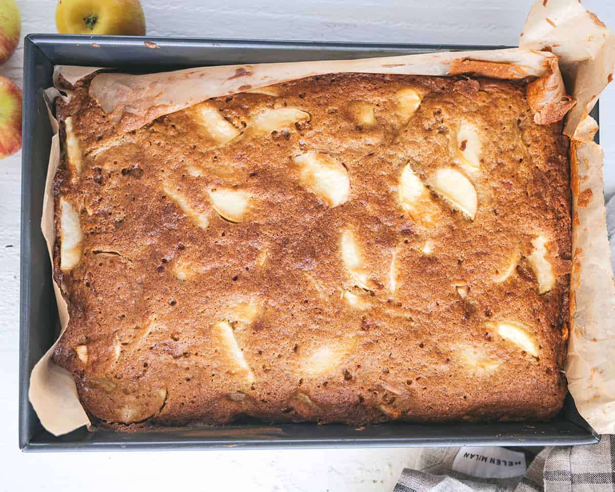 Baked apple cinnamon cake that is golden brown showing apple slices mixed in, top view in the pan. 
