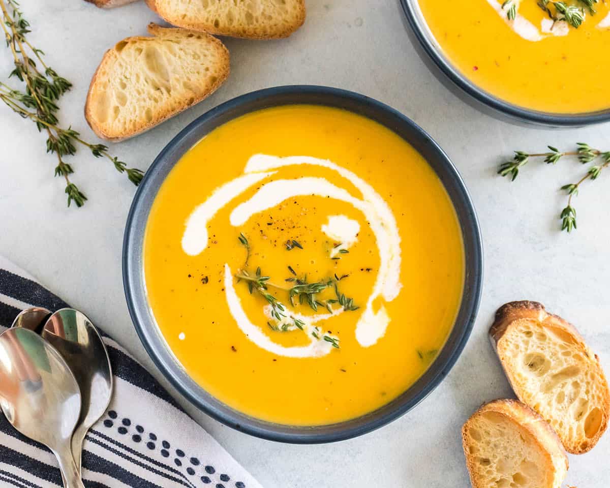 A bowl of butternut squash soup topped with a drizzle of cream and spices, surrounded by bread slices, fresh thyme, and a napkin and spoon. Top view.