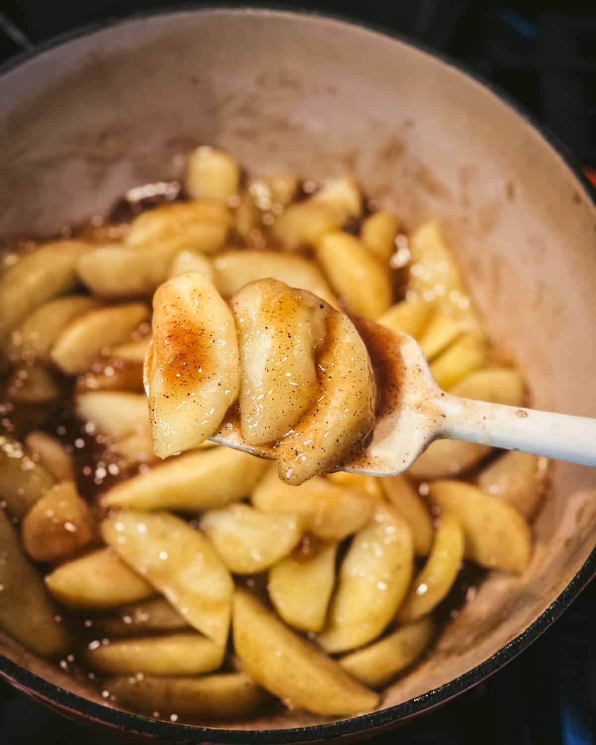 Apple slices cooking with the spices and liquid, reducing down to apple pie filling. A spoon lifting some of the slices up showing their spice and sugar, top view. 