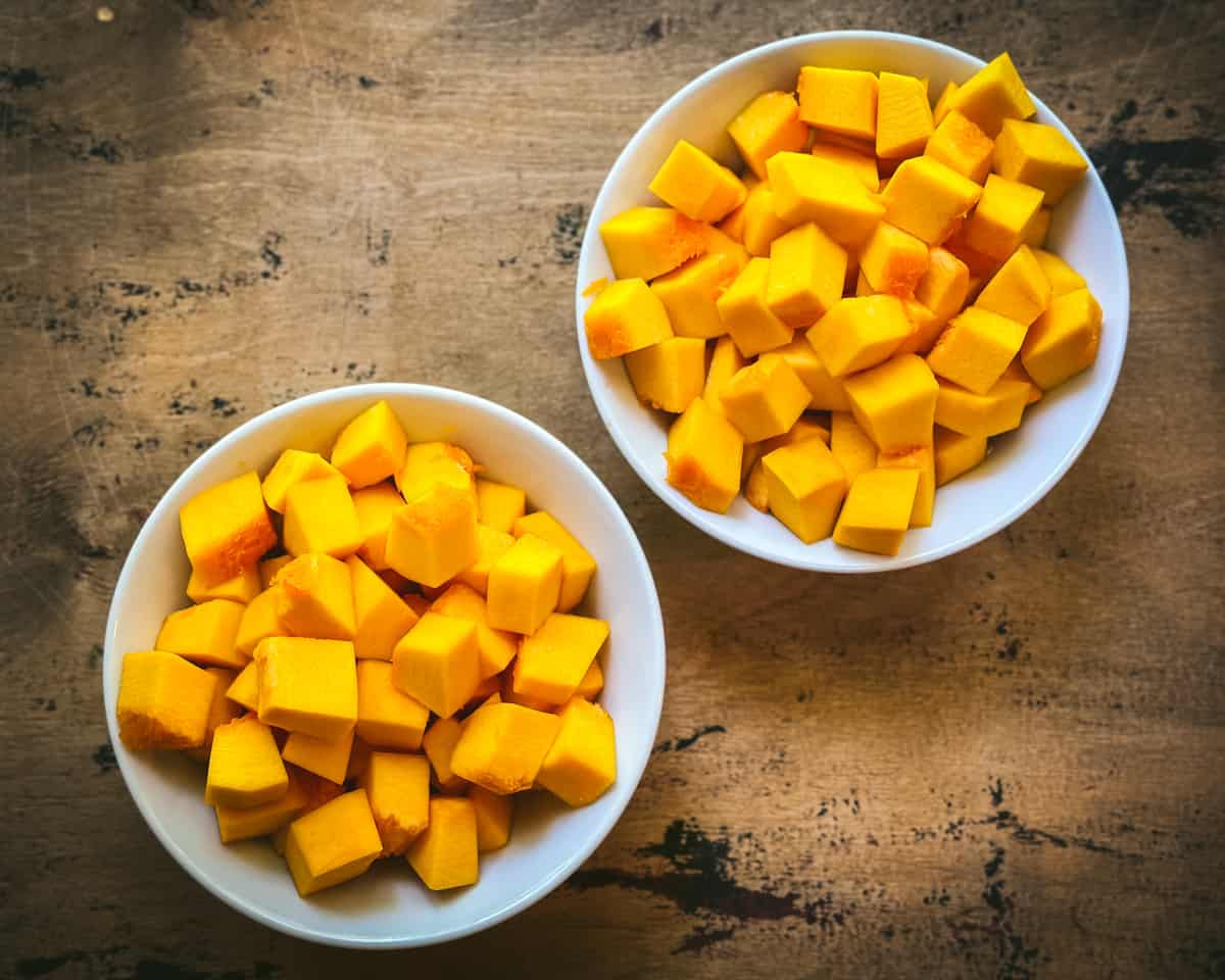 Fresh cubed pumpkin separated into two white bowls on a dark wood surface. 