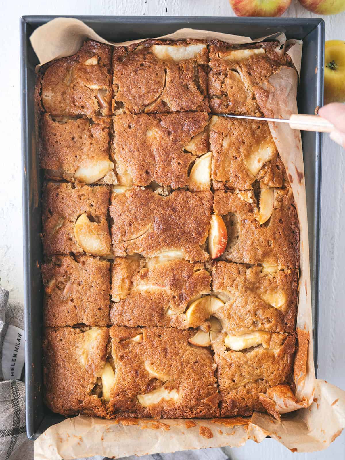 A baked apple cake in a parchment lined pan being cut into squares.