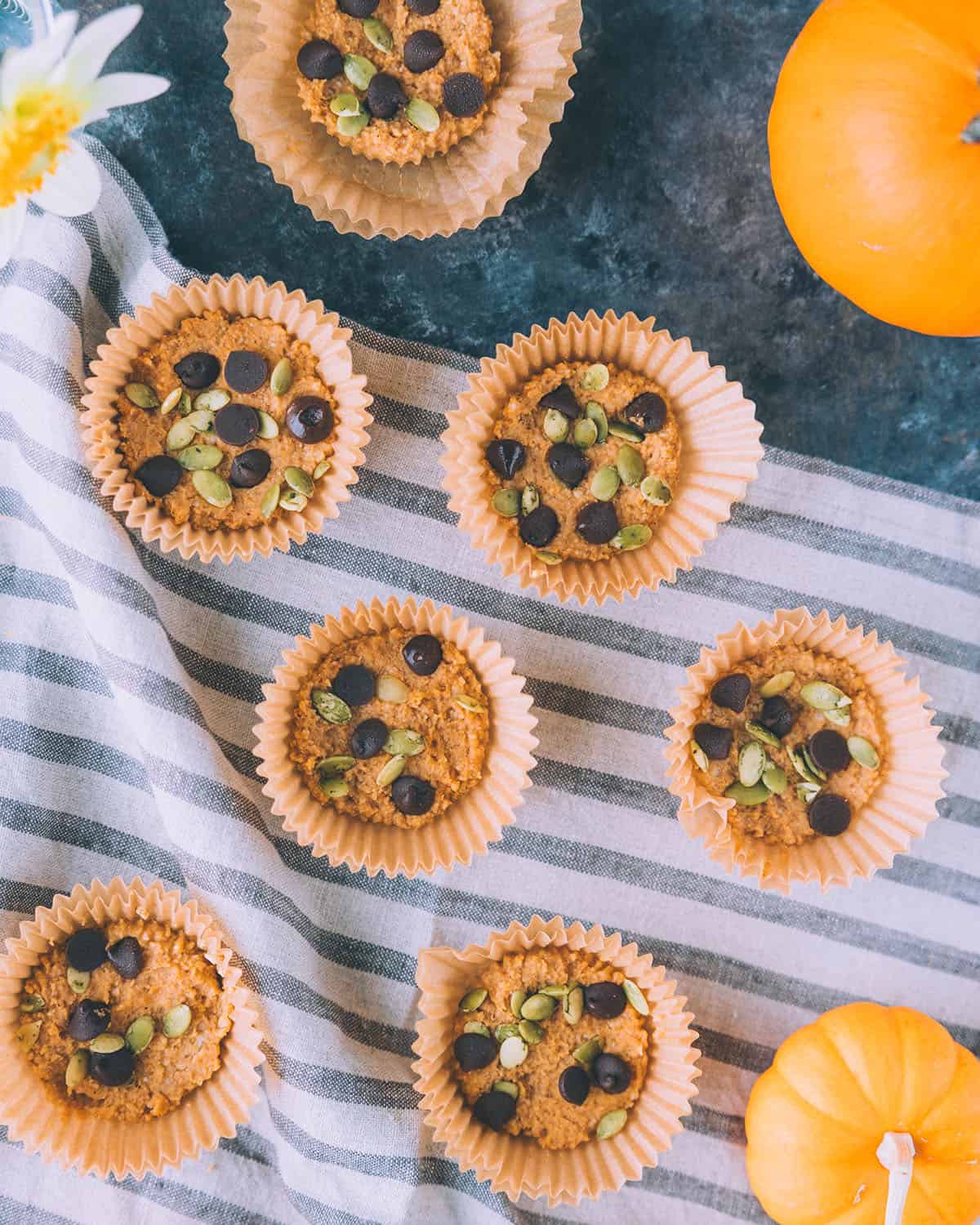 Finished pumpkin cups in papers topped with chocolate chips and pumpkin seeds, on a striped towel surrounded by fresh pumpkins and a white daisy. 