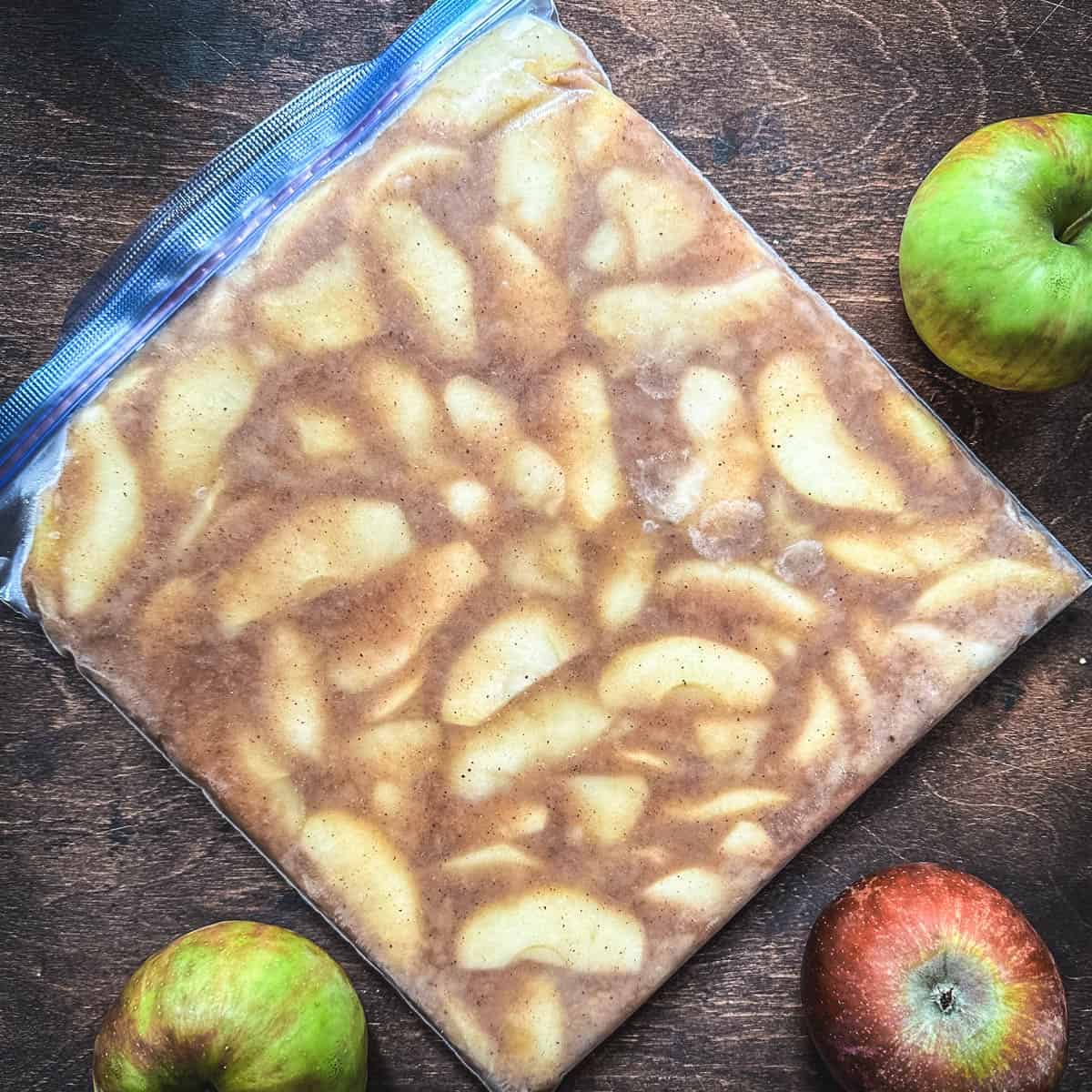 A bag of frozen apple pie filling on a wood surface surrounded by fresh whole apples. Top View.
