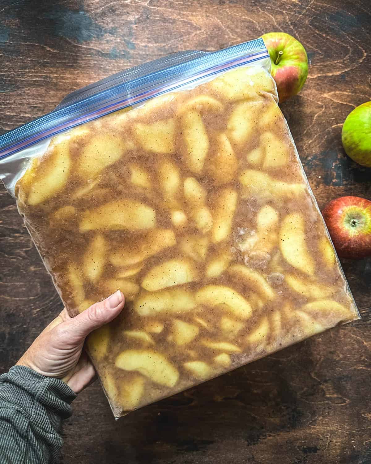 Apple pie filling in a gallon zip top bag being held by a hand over a dark wood surface with fresh apples surrounding. 