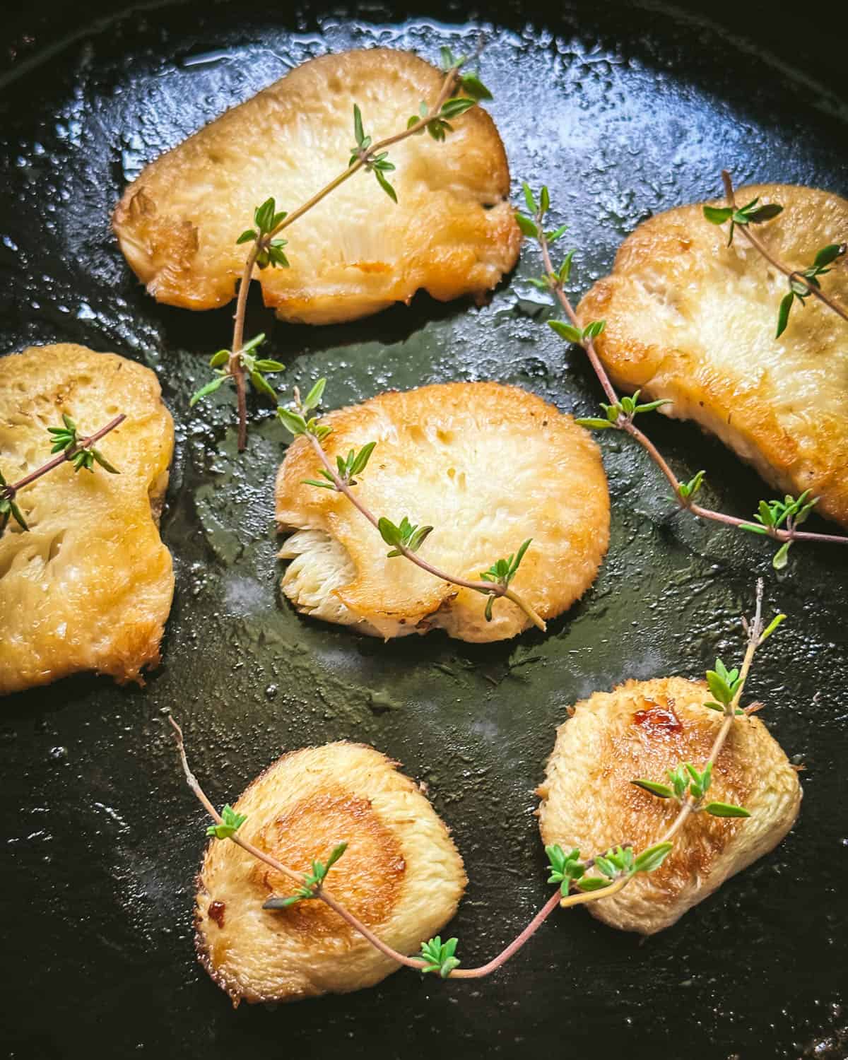 Several slices of lion's mane mushroom golden brown sautéing on a cast iron pan, with fresh thyme sprigs. 