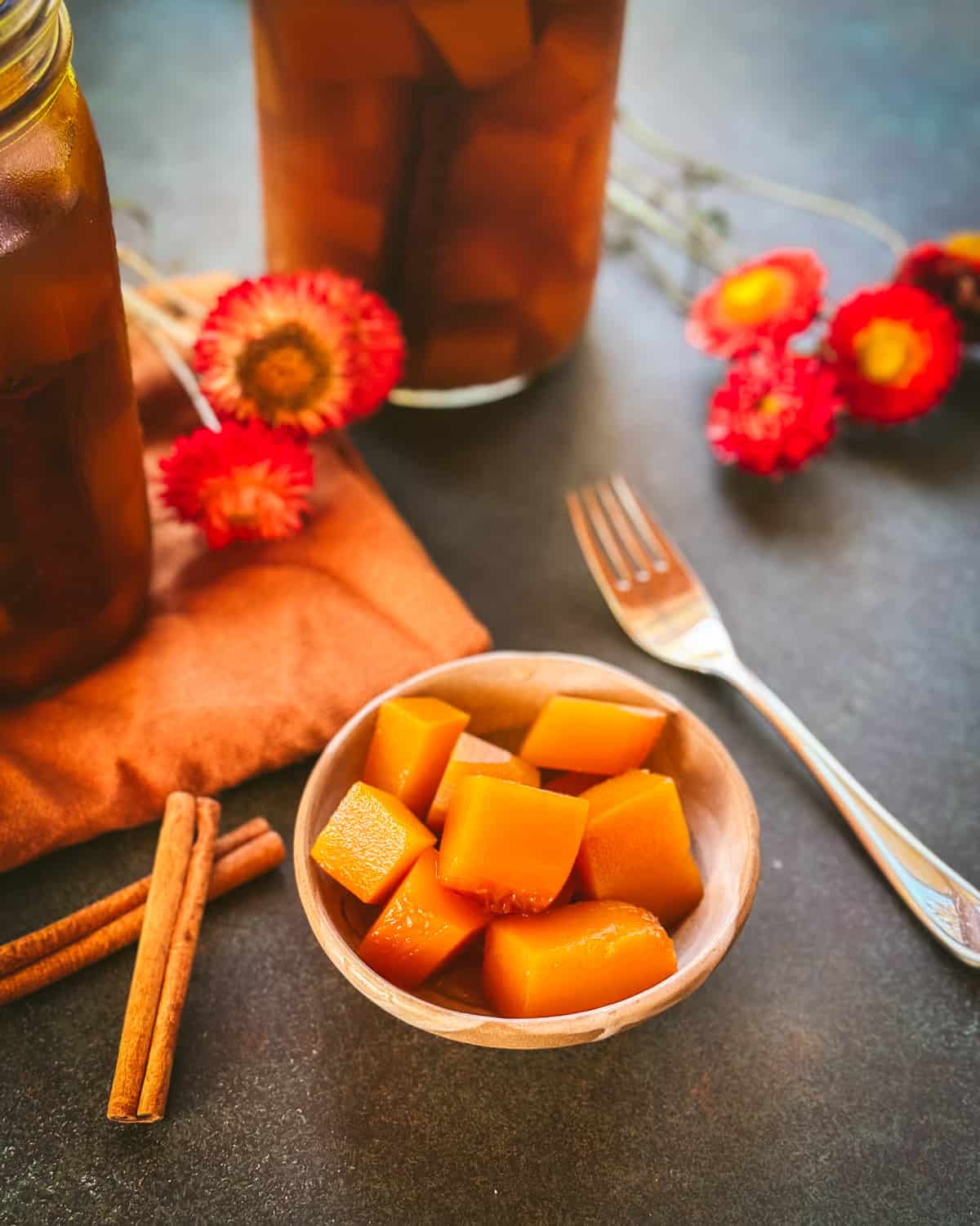 A small white bowl of pickled pumpkin cubes, surrounded by a fork, dried flowers, cinnamon sticks, and jars of pickled pumpkin.