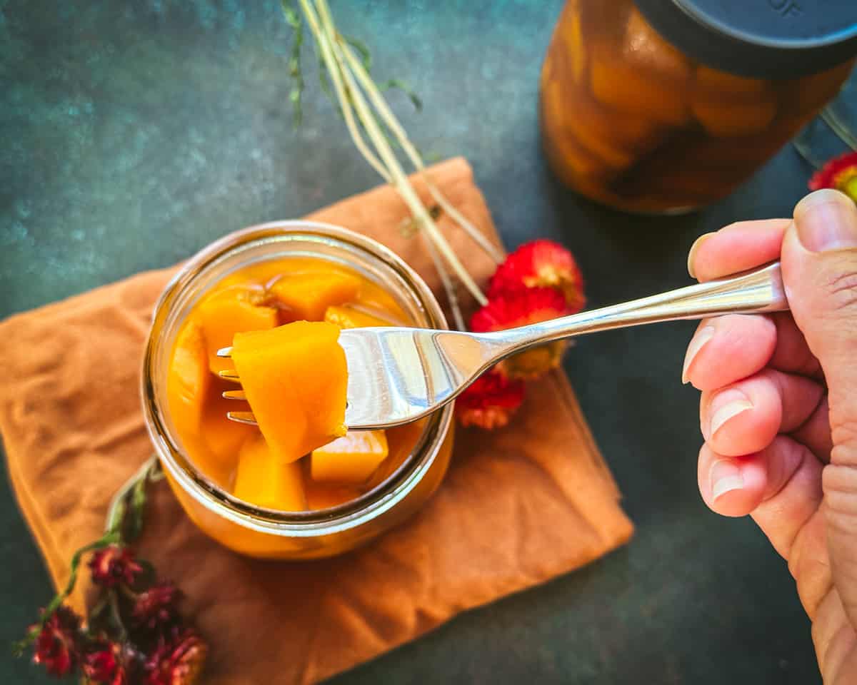 A jar of pickled pumpkins with a fork lifting one up to see close up. On an orange cloth napkin surrounded by flowers and another jar of pumpkin pickles. Top view. 