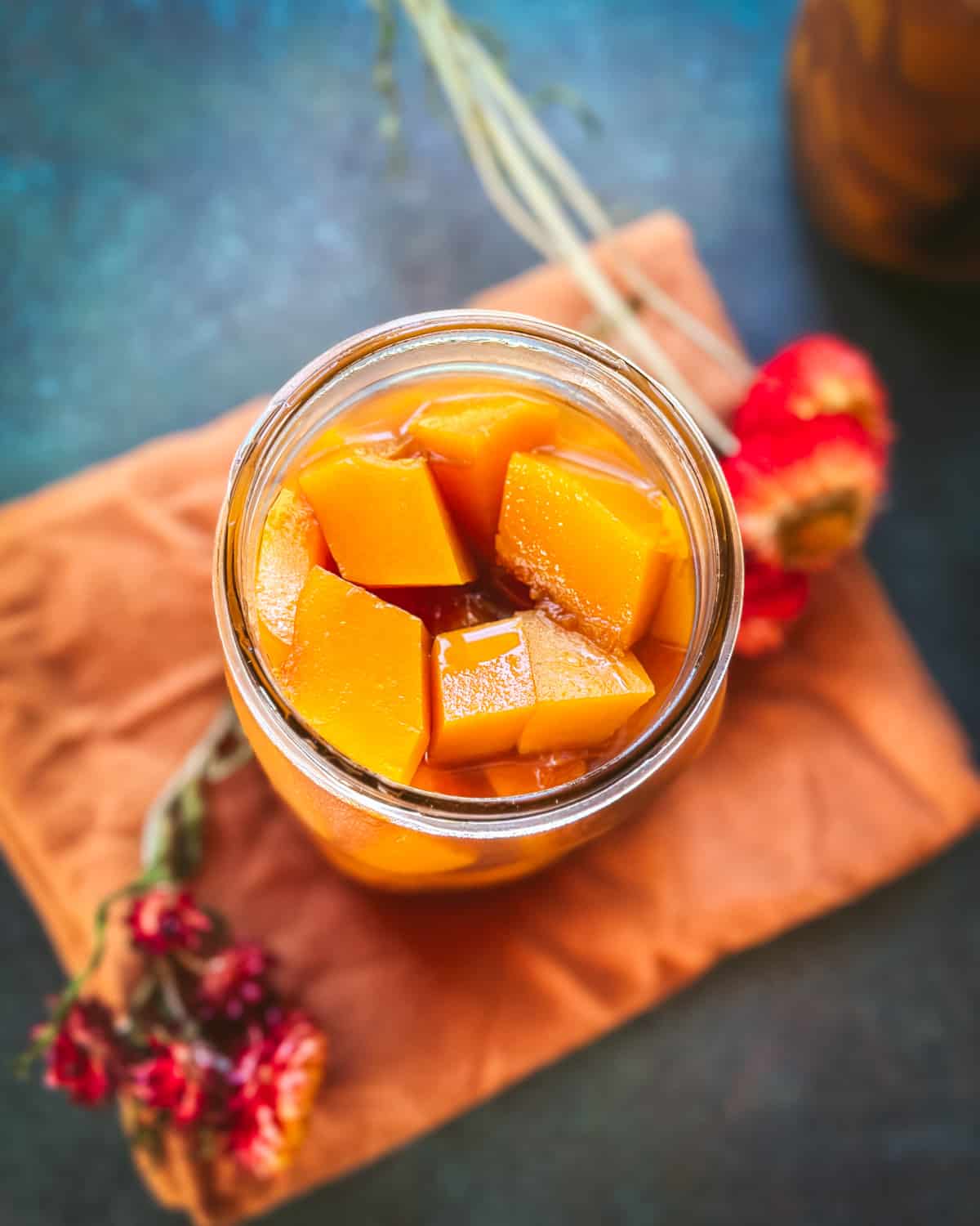 Pickled pumpkin in a jar on an orange cloth napkin, with dried flowers surrounding. Top view. 