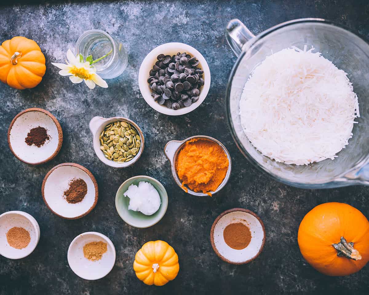 Ingredients for coconut butter pumpkin cups in small bowls on a dark blue countertop, with fresh pumpkins surrounding. 