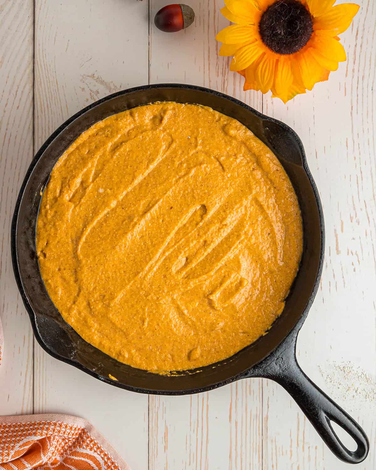A cast iron skillet with pumpkin cornbread batter in it on a white wood surface. 