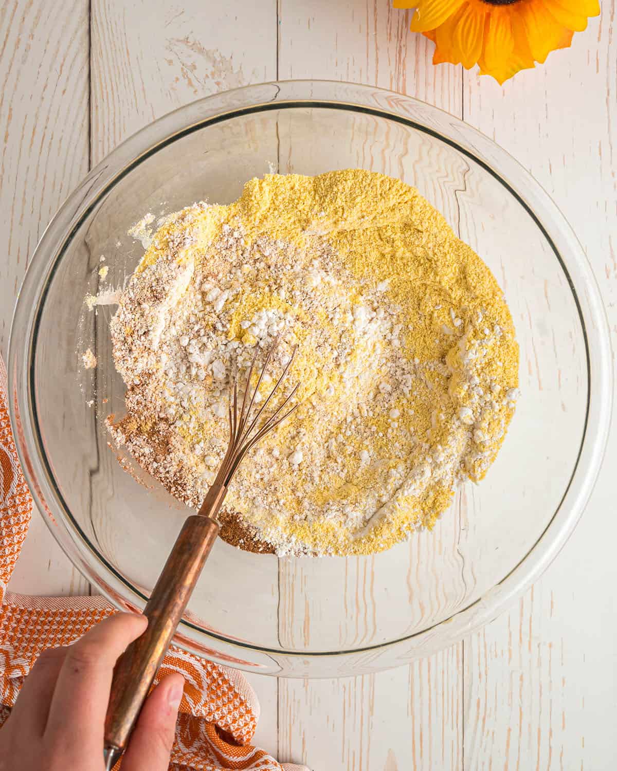 Dry cornbread ingredients in being whisked together in a clear bowl, top view. 